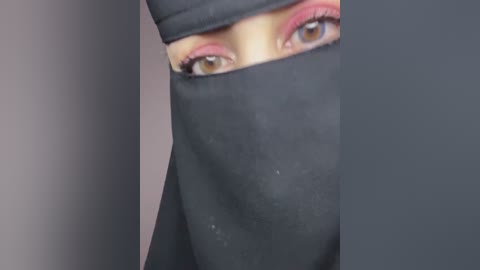 A close-up video of a person's face, partially covered by a black niqab. The individual has light skin and brown eyes, with pink eyeshadow and mascara. The background is a gradient of dark and light gray.