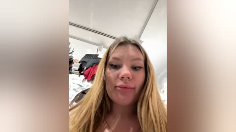 A selfie of a young woman with long blonde hair, wearing a red top, in a modern, sparsely furnished room with white walls and a mirror reflecting cluttered clothes.