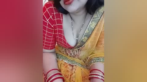 Video of a woman with pale skin and dark hair, wearing a vibrant yellow sari with red borders and intricate gold embroidery, adorned with multiple red bangles and a silver necklace.