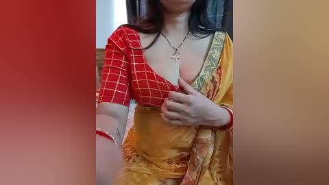Video of a woman in a red and yellow traditional Indian saree with a plunging neckline, revealing ample cleavage, and a cross necklace. She has long dark hair and wears a red bracelet on her left wrist.
