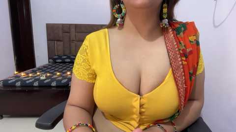 Video of a curvy South Asian woman in a yellow crop top, exposing cleavage, with orange and green sari draped over her shoulder. She wears large, colorful earrings. Background shows a bed with a patterned blanket and fairy lights.