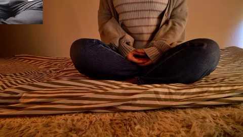 Video of a person sitting cross-legged on a striped bedspread, wearing a beige cardigan and a striped sweater, with a serene expression.