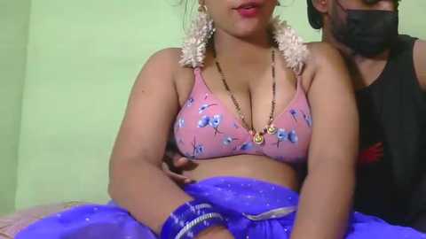 A video of a woman in a pink floral bra and blue saree, wearing a mask, with a man's arm around her, set against a pale green wall.