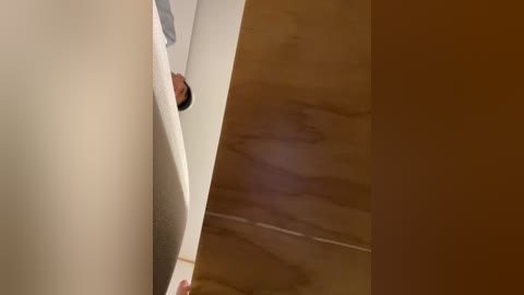 Video of a man in a white shirt peering around a corner into a dimly lit room with a wooden floor, creating a sense of suspense.