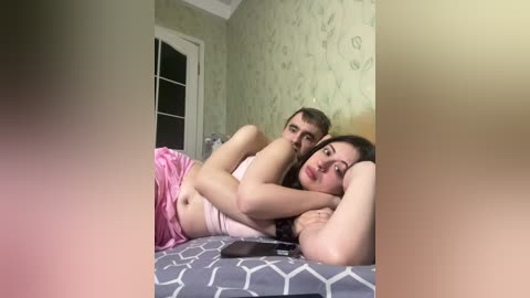 Media: A video of a young couple in bed, with a woman in pink pajamas and a man in a white shirt, both lying on a gray geometric-patterned bedspread. The room has green wallpaper and a partially visible white door.