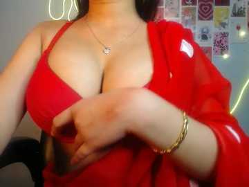 Video of a woman with medium skin tone wearing a red bra and sheer red blouse, partially covering her cleavage. Her hands are on her breasts. Background includes a wall with colorful photos and neon lights.