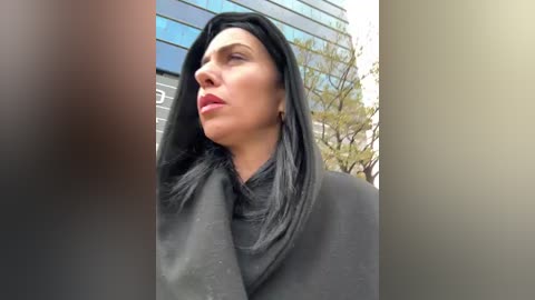 Video of a woman with long black hair, wearing a black hijab, standing outdoors in front of a modern building with reflective windows.