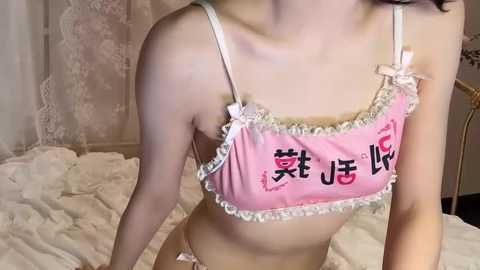 Video of a slender, light-skinned Asian woman in a pink, frilled lingerie set with Japanese characters on the bra. She kneels on a bed with white sheets in a cozy, softly lit room.