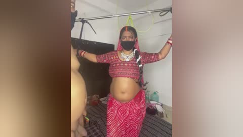 Video of a pregnant woman in traditional red sari, wearing a black mask, standing with arms raised in a small, cluttered room with white walls and a bed.