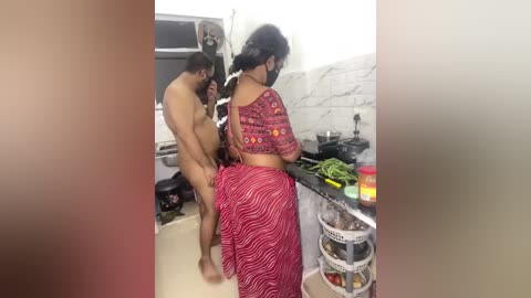 Video of a nude man and woman engaged in a sexual act in a kitchen. The woman is dressed in a red and white striped sari, standing behind the man.