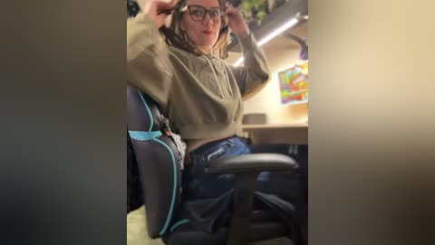 Video of a woman in a gaming chair, adjusting headphones, wearing a green hoodie, glasses, and blue jeans. Background shows a cozy, brightly lit room with a colorful painting and a desk.