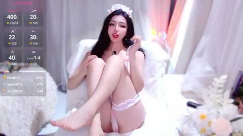 Video of an East Asian woman with long black hair, wearing a white lace teddy and headband, sitting on a white bed, surrounded by soft lighting and fluffy pillows.
