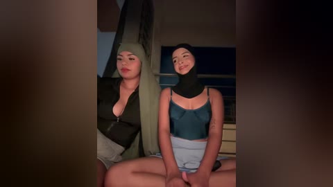 Video of two young women with medium skin tones and dark hair, both wearing hijabs and revealing outfits, seated indoors with a dimly lit background.