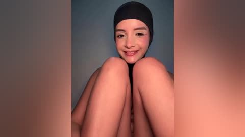 Video of a smiling, light-skinned woman with medium-length black hair and dark eyes, wearing a black swim cap, sitting with legs bent and knees touching, against a muted blue-gray background.