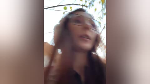 A blurry video of a woman with long brown hair, wearing glasses, and a dark top, standing outdoors with green foliage in the background.