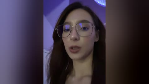 Video of a young Caucasian woman with fair skin, long brown hair, and glasses, slightly blurred, in a dimly lit room with purple and white lighting.