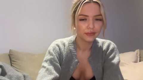 A video of a blonde woman with fair skin, wearing a grey cardigan, and a low-cut black top, sitting on a beige couch. She has a slight smile and is in a living room with light-colored walls.