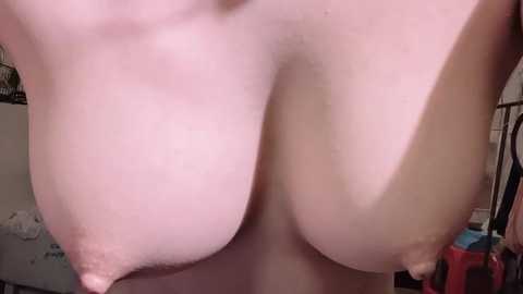 Video of a fair-skinned, topless woman with large, natural breasts, standing indoors. Background includes a white dresser and a red bucket, suggesting a domestic setting.