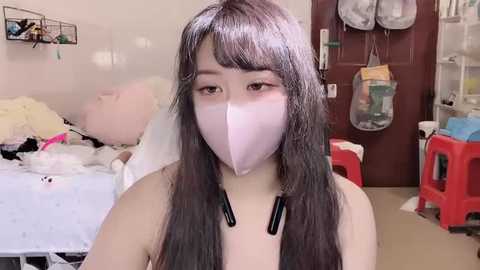 Video of an East Asian woman with long black hair, wearing a white face mask, standing in a cluttered bedroom with a messy bed, red plastic chair, and hanging laundry.