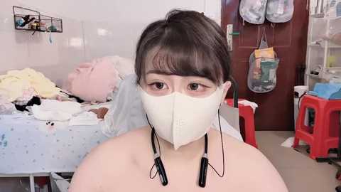 Video of an Asian woman with short black hair, wearing a white surgical mask, sitting in a cluttered hospital room with medical equipment, laundry, and a bed in the background.
