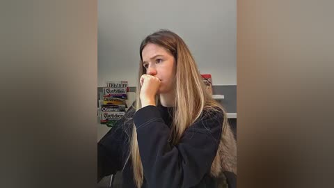Video of a young Caucasian woman with long, blonde hair, wearing a black jacket, standing pensively in a room with a gray wall and a colorful bookshelf behind her.