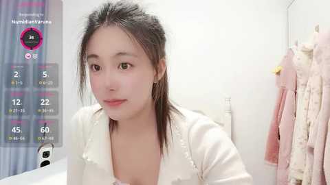 A young Asian woman with light skin and straight black hair, wearing a white top, stands in a bathroom with a pink towel hanging on the wall.