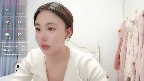 Video of an Asian woman with light skin, dark hair, and a white robe, looking thoughtful. Background features a bed with pink and white bedding and a blue curtain. A digital overlay shows live stream statistics.