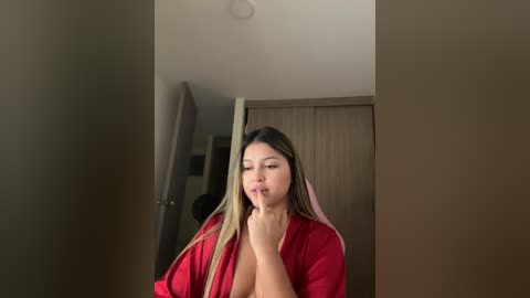 A video of a young woman with long, straight, blonde hair, wearing a red robe, standing in a doorway with her finger to her lips.