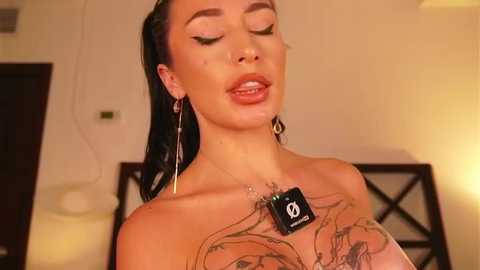Video of a fair-skinned woman with long, dark hair, wearing a black strapless top, showcasing extensive tattoos on her chest and arms. She has striking makeup, including dark eyeliner and nude lipstick, and wears dangling earrings. Background includes a beige wall and a bed with a white lamp.