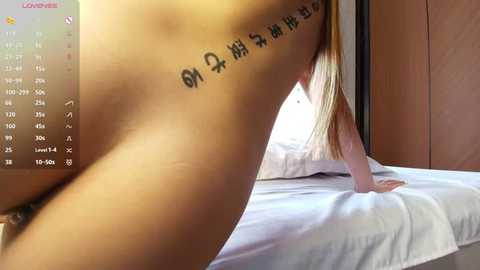 Video of a nude Asian woman with long blonde hair, kneeling on a bed with white sheets, showing a tattooed Chinese character on her side.