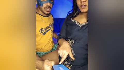 Video of a man and woman in a dimly lit room. The man wears a yellow shirt and a blue mask, while the woman has dark hair and a navy blue outfit.