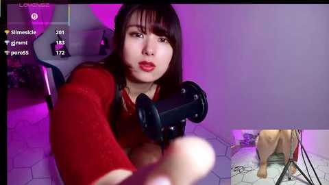 Video of a woman with long dark hair, red lipstick, and a red sweater, using a microphone in a purple-lit room.
