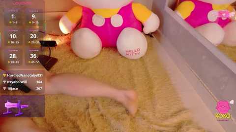 Video of a plush Hello Kitty doll with a pink, yellow, and white outfit, positioned on a beige, fluffy rug. A pink vibrator is partially visible. The background includes a mirror reflecting the doll.