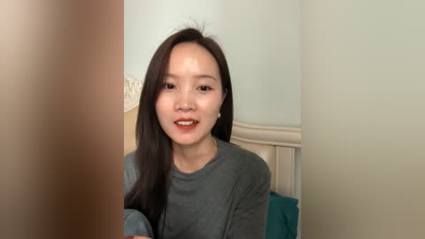 Video of an East Asian woman with long black hair, wearing a gray sweater, sitting on a bed with light beige walls and teal pillows.