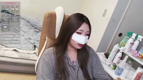 Video of a woman in a gray striped shirt, wearing a white surgical mask, sitting on a tan leather office chair, with a messy desk behind her.