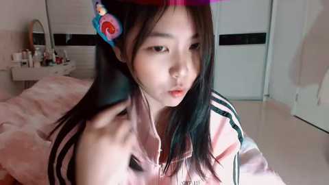 Video of a young Asian woman with long black hair and a red flower headband, wearing a pink and white striped hoodie, sitting on a bed in a bedroom with a mirror, shelves, and a closet in the background.