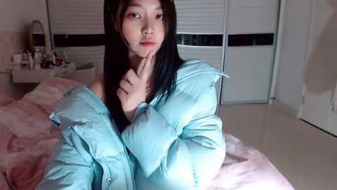 Video of an Asian woman with long black hair, wearing a light blue puffy jacket, sitting on a bed in a dimly lit bedroom with white walls and a closed closet.