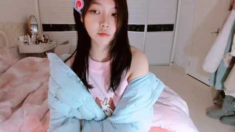 A video of an Asian woman with long black hair and a pink flower in her hair, wearing a light pink shirt and a blue blanket, in a cozy bedroom with white walls and a vanity table.