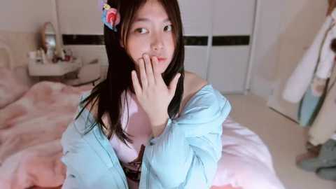 Video of an Asian woman with long black hair and a pink hair accessory, wearing a light blue off-shoulder blouse, sitting on a pink bed in a softly lit bedroom.
