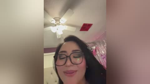 Video of a smiling woman with long dark hair, wearing black-framed glasses, standing in a pink-painted room with a ceiling fan.