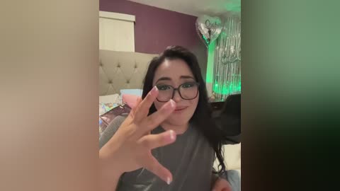 Video of a young woman with long black hair, glasses, and fair skin, wearing a grey top, standing in a dimly lit room with green fairy lights, a bed, and a chair.
