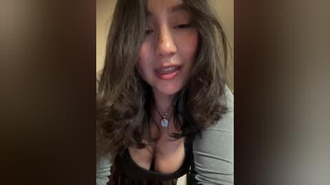 A close-up video of a smiling East Asian woman with shoulder-length, wavy black hair and a medium complexion, wearing a black top that reveals cleavage. She has a silver necklace with a pendant. The background is blurred.