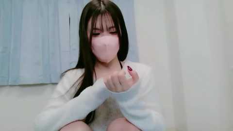 A video of an East Asian woman with long black hair and fair skin, wearing a white sweater and pink mask, sitting on a bed, holding a bottle, with light blue curtains in the background.