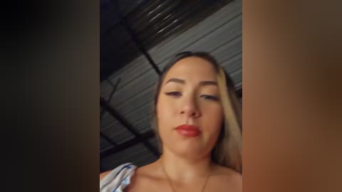 A video of a young woman with fair skin, straight blonde hair, and red lipstick, wearing a light blue shirt, standing in a dimly lit warehouse with metal beams and a corrugated roof.