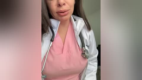 A video of a light-skinned woman with shoulder-length brown hair, wearing a pink scrub top and white lab coat, holding a stethoscope. The blurred background suggests an indoor medical setting.