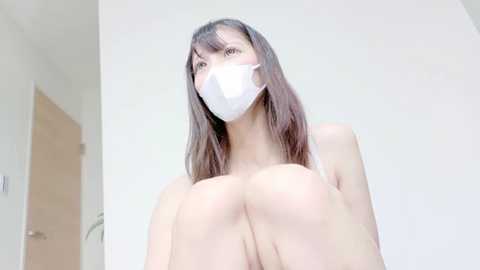 Video of a young East Asian woman with long, straight brown hair, wearing a white surgical mask, sitting on a bed, looking up, in a minimalist white room.