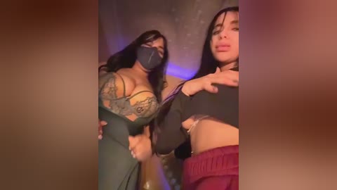 Video of two women in a dimly lit room, one with large breasts and tattoos, wearing a black mask, and the other with long black hair and a black top, both with a playful, confident expression.