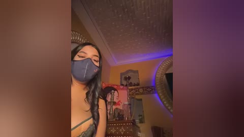 Video of a woman with long black hair, wearing a blue face mask and a revealing blue lace bra, in a dimly-lit room with decorative items, including a round mirror, and a wall with posters.