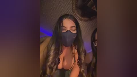 Video of a woman with long, wavy black hair, wearing a black face mask, a low-cut top revealing cleavage, and a beige jacket, sitting on a chair in a dimly lit room.