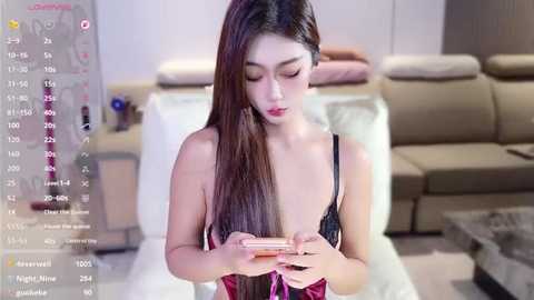 A video of an Asian woman with long, straight black hair, wearing a black lace bra, sitting in a modern living room, focused on her smartphone.